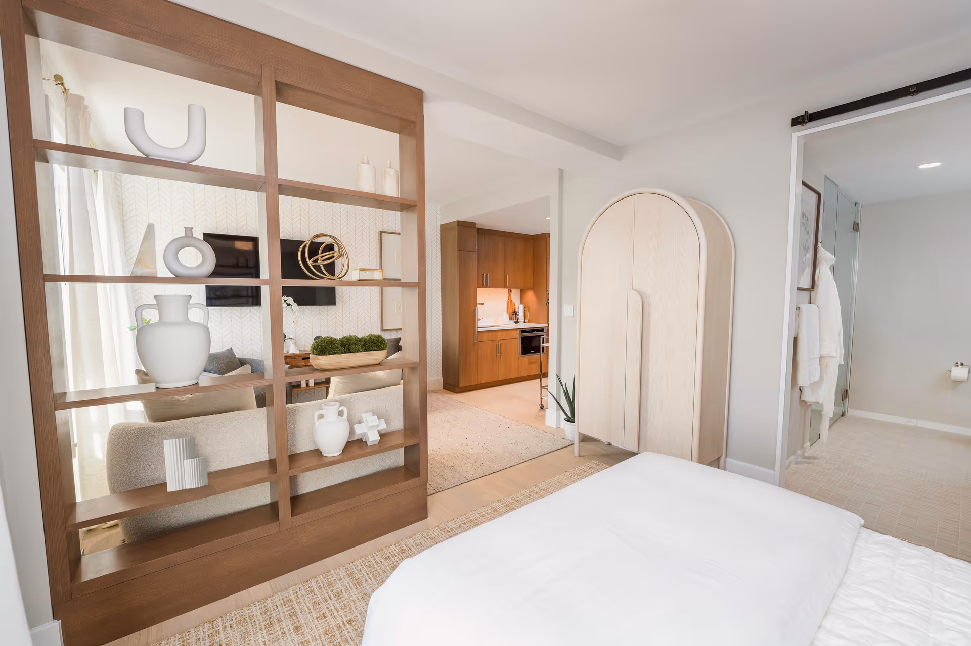A modern, bright bedroom with a white bedspread in the foreground. A wooden open shelving unit with decorative items separates the bedroom from a living area with a wall-mounted TV and a sofa. In the background, there is a kitchen area with wooden cabinets and a small dining table. To the right, a sliding door leads to a bathroom with a glass shower and a white robe hanging on the wall.