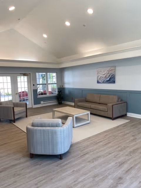 A bright and spacious living room with vaulted ceilings, recessed lighting, light wood flooring, and a large area rug. The room features a beige sofa, two armchairs, a rectangular coffee table, a potted plant in the corner, and a piece of abstract wall art above the sofa. French doors and windows allow natural light to enter the space.