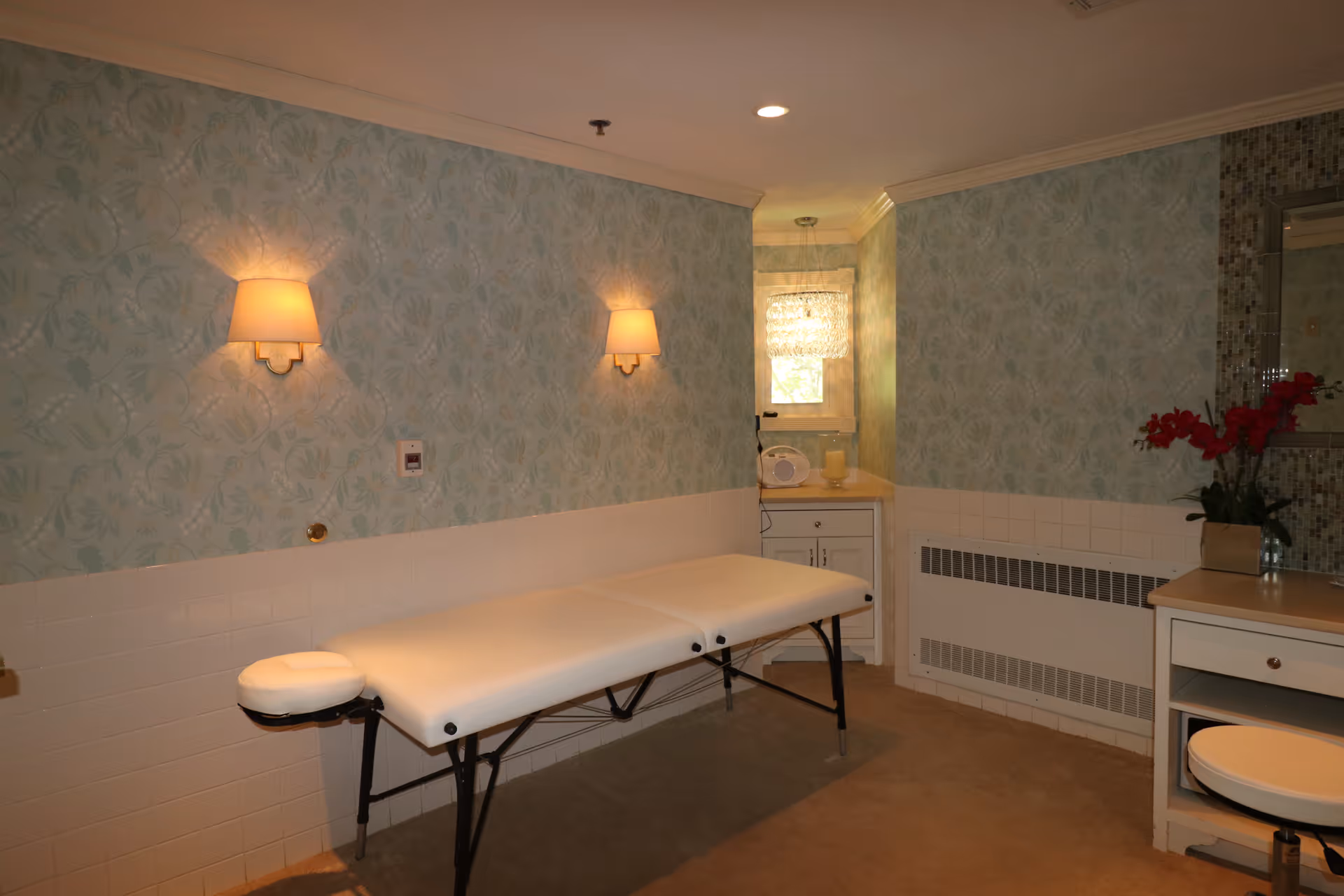 A treatment room featuring a white massage table, floral wallpaper, wall sconces, a small sink cabinet, and a vase of red flowers.