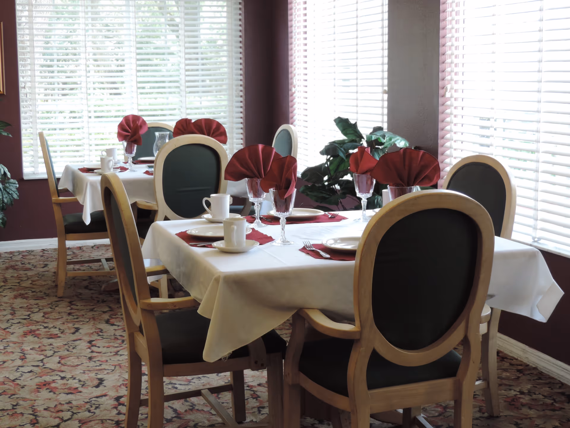 A dining room with two tables set for four people each. The tables have white tablecloths, red folded napkins in glasses, white plates, cups, and silverware. The chairs have wooden frames with dark green cushions. Large windows with white blinds let in natural light, and there is a green plant in the corner.