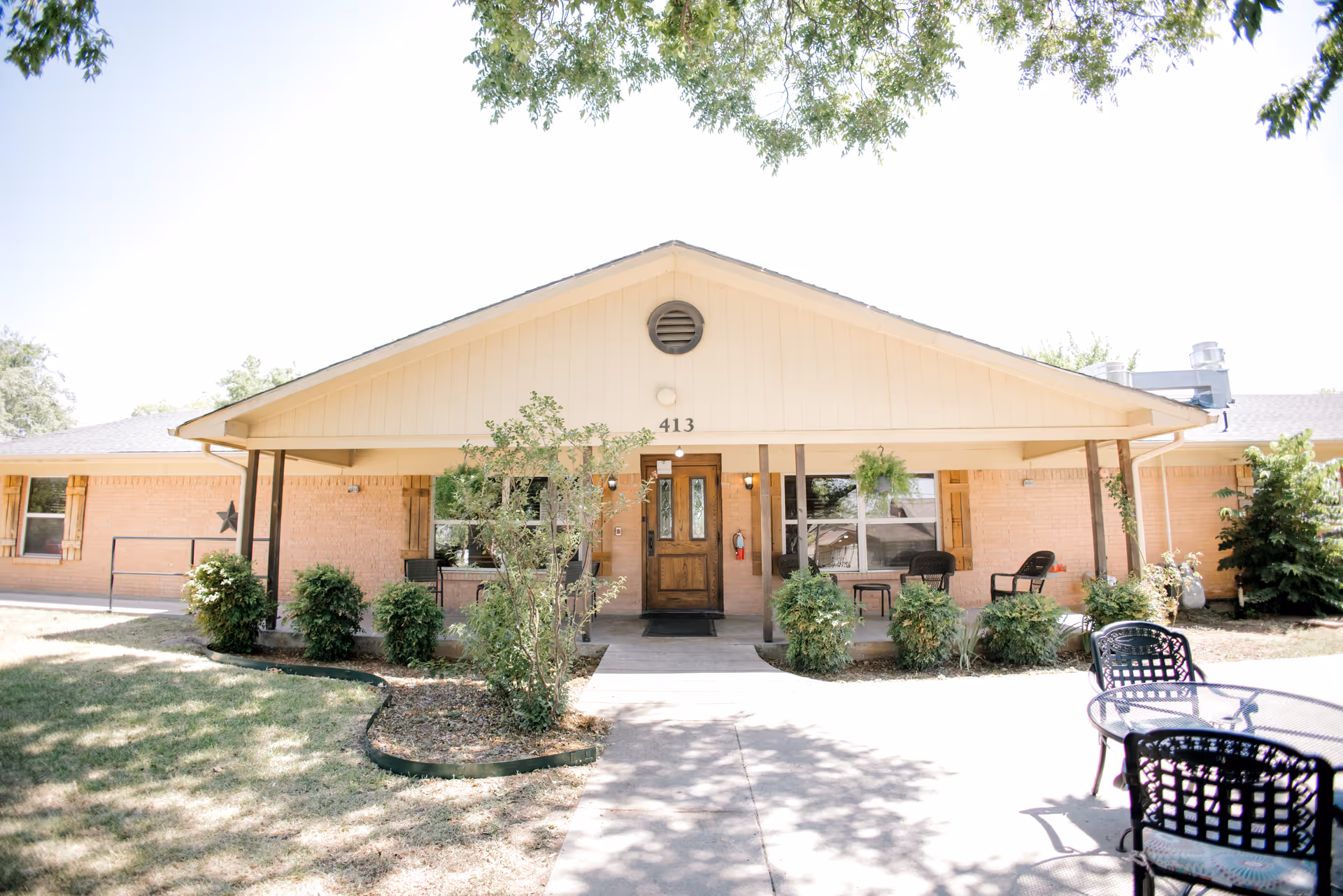 Front entrance of a single-story brick senior living facility with a covered porch, shrubs, and outdoor seating.