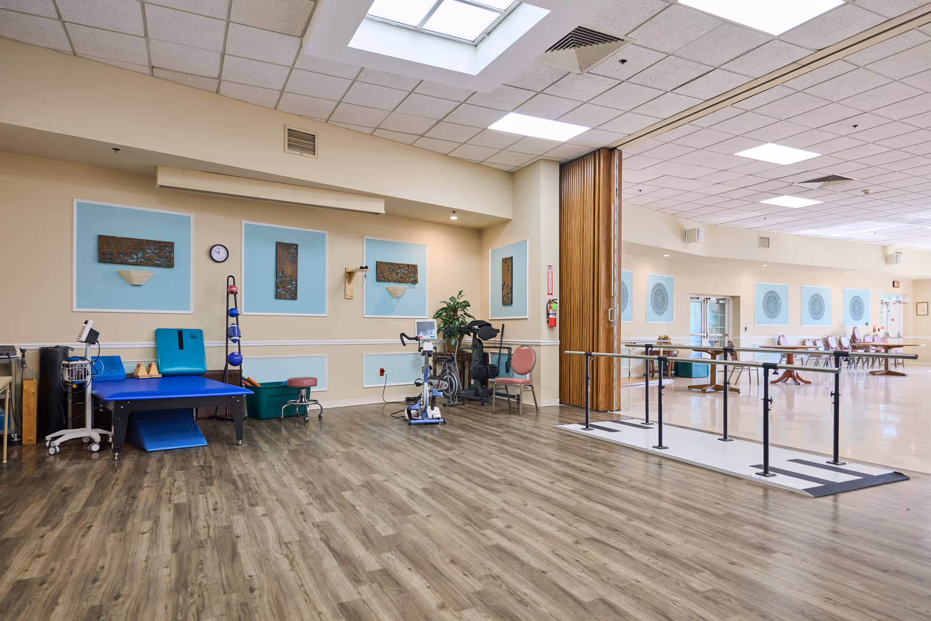 A spacious rehabilitation room with wooden flooring, exercise equipment including a therapy table, stationary bike, and parallel bars for walking practice. The walls are decorated with framed artwork and light blue panels. The room is well-lit with ceiling lights and a skylight, and there is a folding partition dividing the space from an adjacent area with tables and chairs.