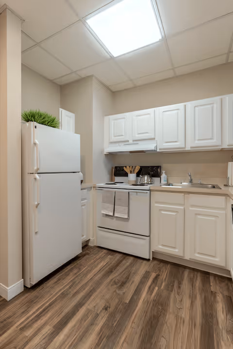 A compact kitchen area with white cabinets, a white refrigerator, a white stove with two towels hanging on the handle, a stainless steel sink, and a countertop with kitchen utensils and a soap dispenser. The floor has wood-patterned vinyl flooring and the ceiling has white tiles with a fluorescent light.