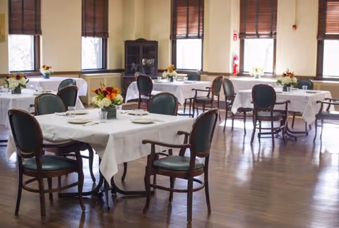 A dining room with several tables covered in white tablecloths, each set with plates, glasses, and floral centerpieces. The room has wooden floors, large windows with brown blinds, and wooden chairs with green cushions around the tables.