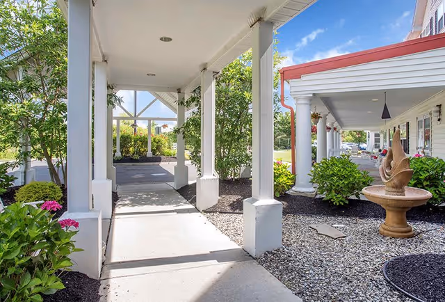 Covered walkway with white columns and a concrete path leading through landscaped garden beds with green shrubs and pink flowers, adjacent to a building with white siding and a red roof edge. A decorative stone fountain is visible on the right side under the covered area.