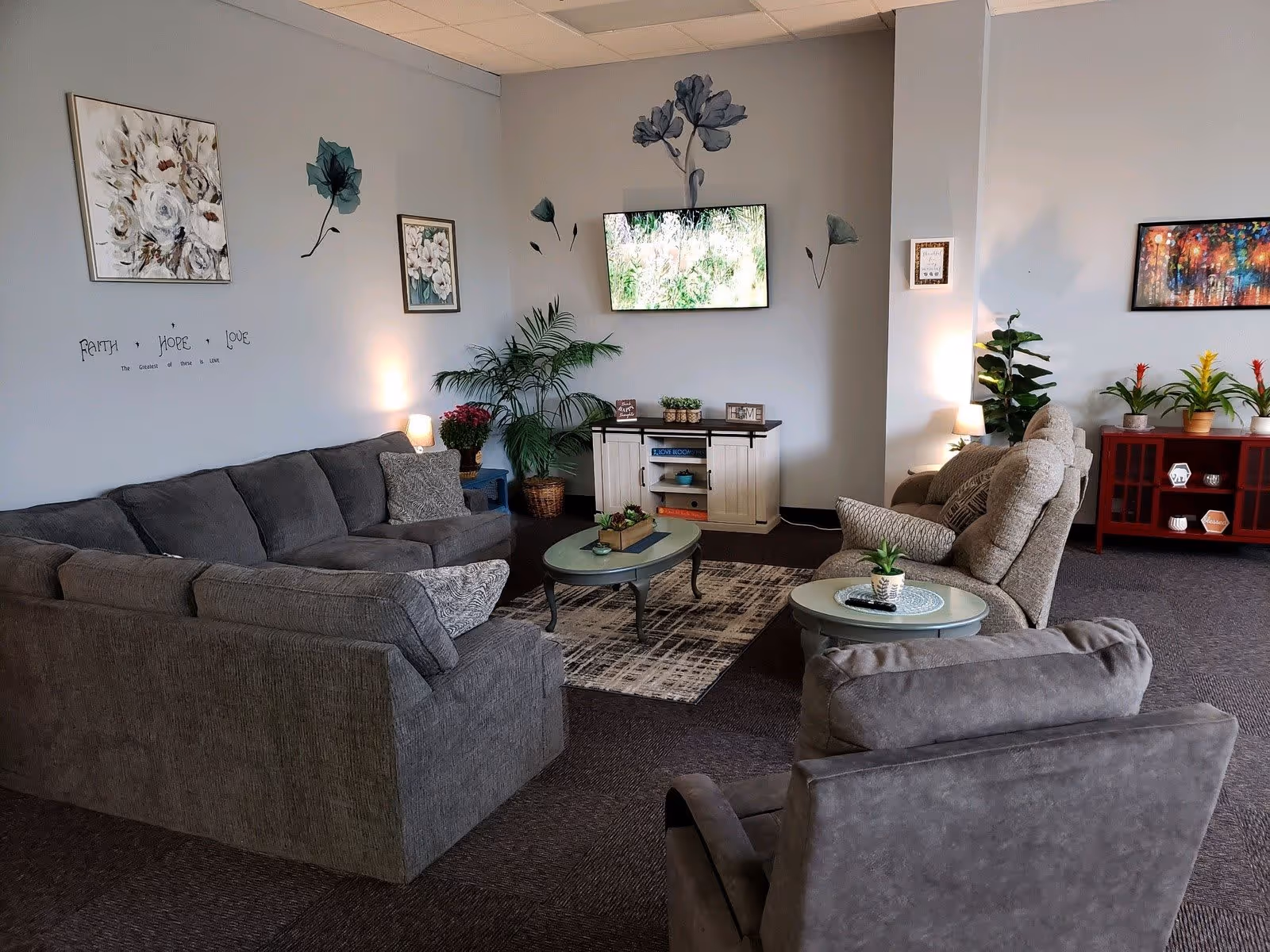 A cozy living room area with a large gray sectional sofa, two gray armchairs, and a small round coffee table with a plant on it. The walls are decorated with floral artwork and wall decals. A flat-screen TV is mounted on the wall above a white cabinet. There are several potted plants and soft lighting from table lamps, creating a warm and inviting atmosphere.
