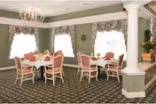 A dining room with several round tables covered with white tablecloths, each surrounded by pink cushioned chairs. The room has green walls with white trim, large windows with floral valances, a chandelier hanging from the ceiling, and a patterned carpeted floor.