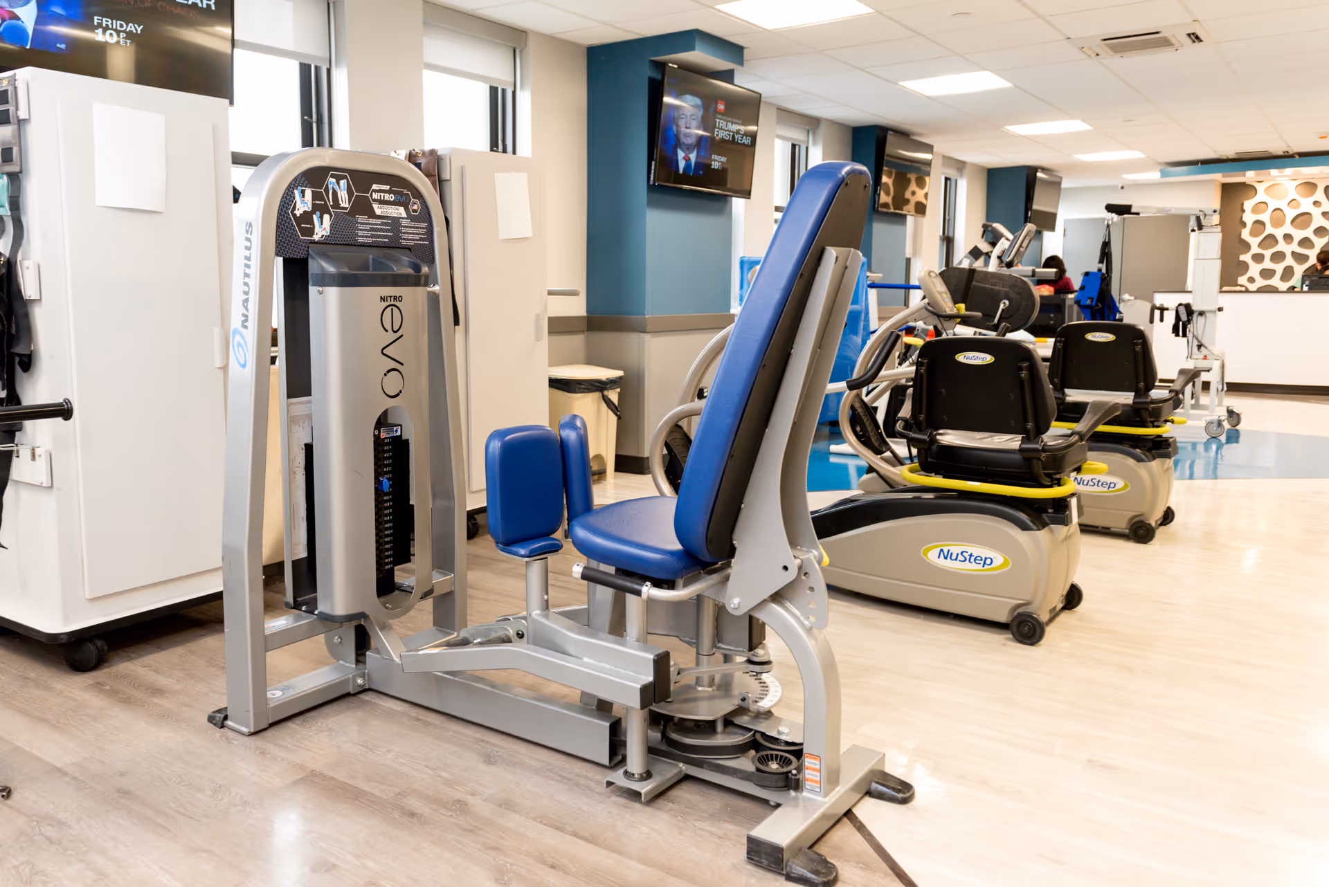 A rehabilitation center room with exercise equipment including a Nautilus Nitro Evo leg press machine and several NuStep recumbent cross trainers. The room has light-colored flooring, blue and white walls, and multiple windows letting in natural light. Two wall-mounted televisions are visible, and there is a reception or check-in desk in the background.