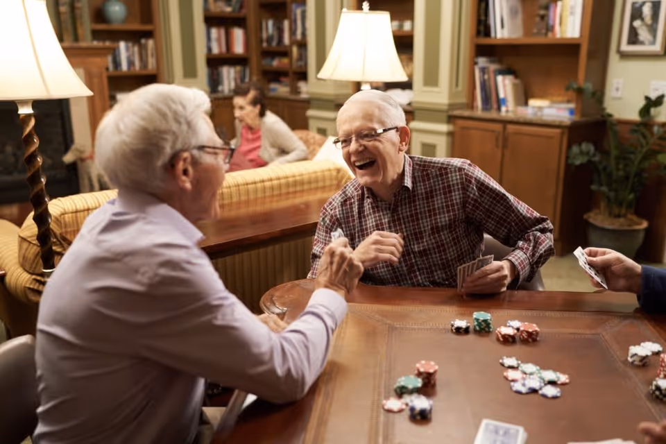 Two elderly men sitting at a wooden table playing cards and poker chips, smiling and enjoying each other's company in a cozy living room with bookshelves, lamps, and a woman sitting on a couch in the background.