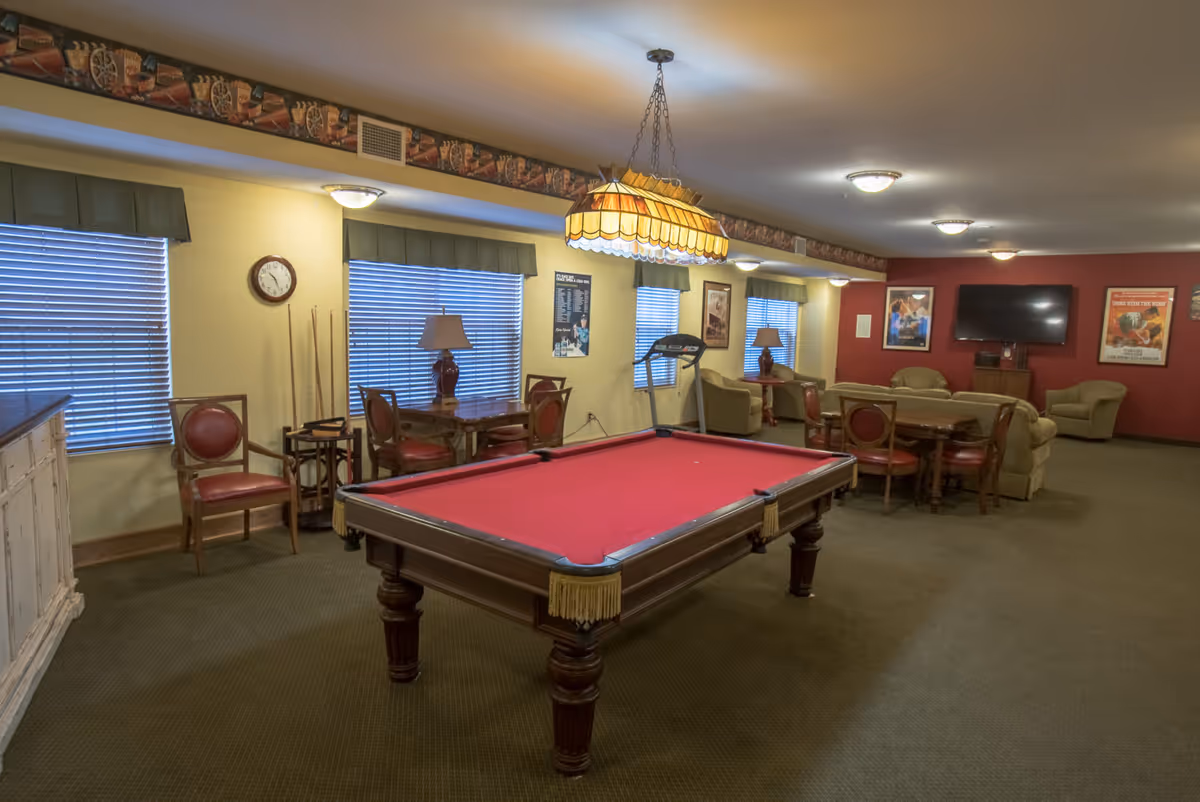 A spacious recreational room with a red felt pool table in the center, surrounded by wooden chairs with red cushions. The room features multiple windows with blinds, a hanging stained glass light fixture above the pool table, several tables with lamps, a treadmill, and a seating area with sofas and armchairs facing a wall-mounted flat-screen TV. The walls are painted yellow and red, decorated with framed posters and a clock.