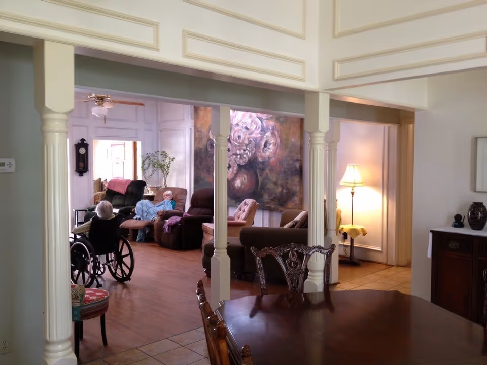 Interior view of a senior living facility common area with elderly residents. One person is seated in a wheelchair, and another is reclining on a sofa. The room features wooden flooring, a large floral painting on the wall, a standing lamp, and classic wooden furniture including a dining table and chairs in the foreground.
