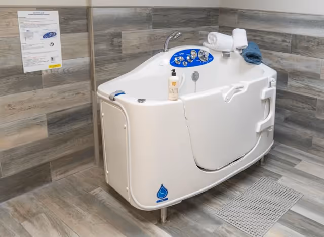 A white walk-in bathtub with control knobs and a faucet, placed in a room with gray wood-patterned tile walls and floor. The bathtub has a door for easy access, a bottle of lotion on the edge, and two rolled towels on top. A drain grate is visible on the floor in front of the tub, and an instruction sheet is mounted on the wall behind it.