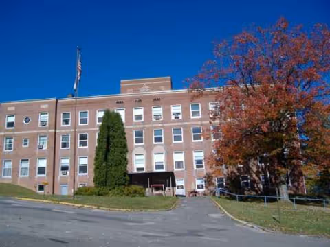 Brick hospital building with numerous windows, flagpole, and autumn trees along a driveway.