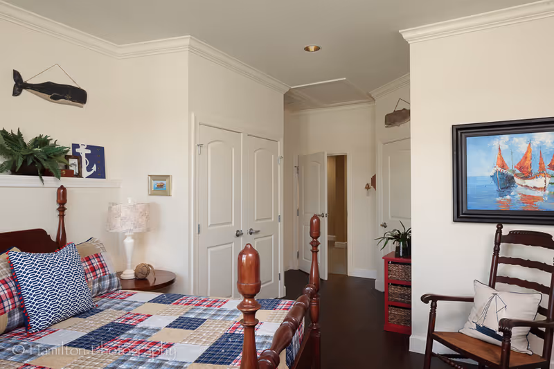 A cozy bedroom with a wooden bed frame and a patchwork quilt in red, blue, and beige tones. There is a bedside table with a lamp and decorative items, a wooden chair with a nautical-themed pillow, and a painting of sailboats on the wall. The room has cream-colored walls, dark wood flooring, and white double doors leading to another area.