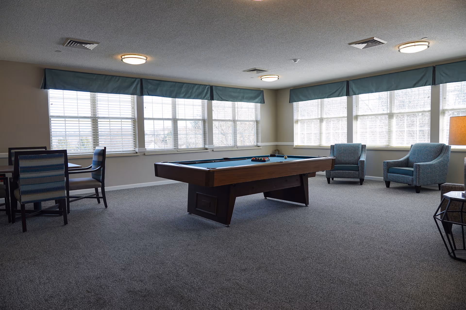 A spacious room with large windows covered by blue valances, featuring a pool table in the center with billiard balls arranged on it. To the left, there is a small table with four chairs, and to the right, two blue upholstered armchairs are positioned near a lamp and a small side table. The room has gray carpet and ceiling lights.
