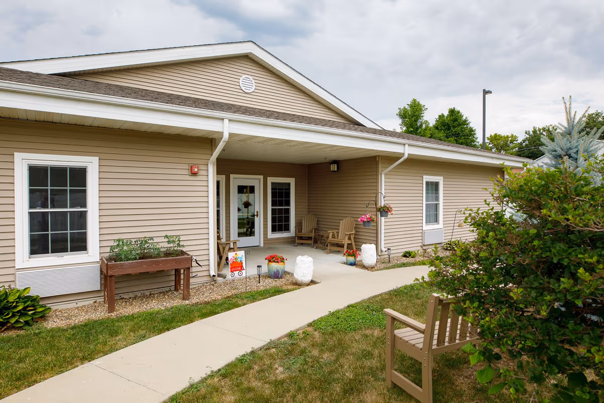 Exterior view of a beige single-story building with white trim, featuring a covered porch area with two wooden chairs and potted plants. A concrete walkway leads to the entrance, and there is greenery including bushes and grass around the building under a cloudy sky.