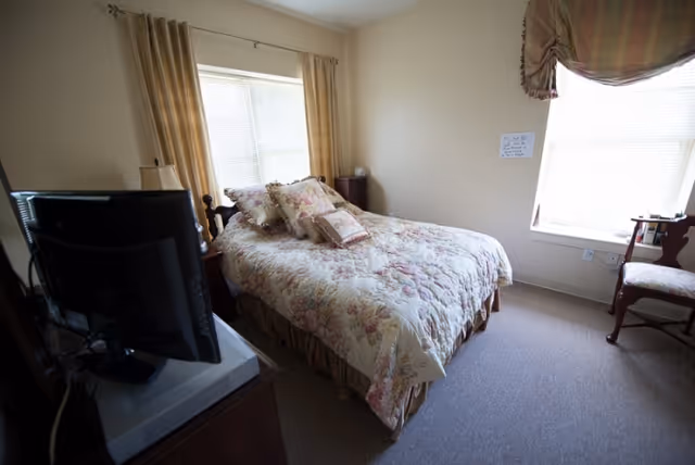 A small bedroom with a bed covered in a floral patterned comforter and multiple pillows. There is a window with beige curtains behind the bed and another window with a valance on the adjacent wall. A wooden chair is placed near the second window, and a television is positioned on a stand opposite the bed.
