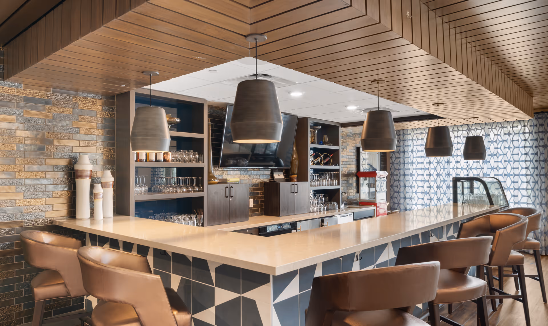 Modern bar area with a long counter featuring geometric patterned tiles, surrounded by brown leather bar stools. The back wall has shelves with glassware and decorative items, a mounted flat-screen TV, and a popcorn machine. The ceiling has wooden paneling with several pendant lights hanging above the counter. A large window with patterned curtains allows natural light into the space.