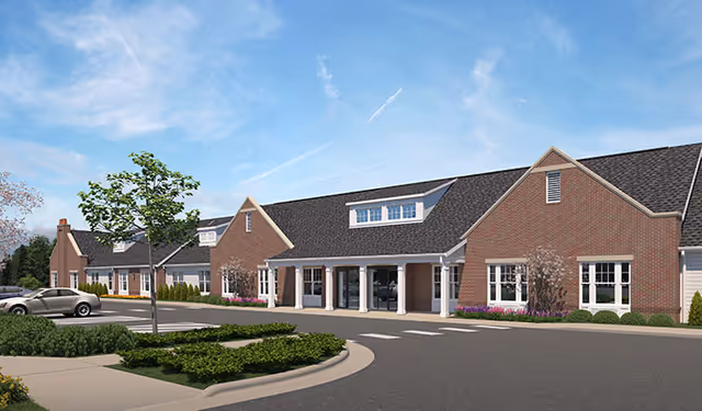 Front exterior of a single-story brick senior living building with a columned entrance, parking area and landscaped islands under a blue sky.