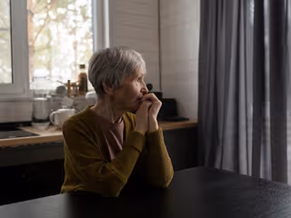 An elderly woman with short gray hair wearing a mustard-colored cardigan sits at a dark table in a dimly lit room, looking thoughtfully out a window with her hands clasped near her face. The background shows kitchen items and a window with light filtering through.