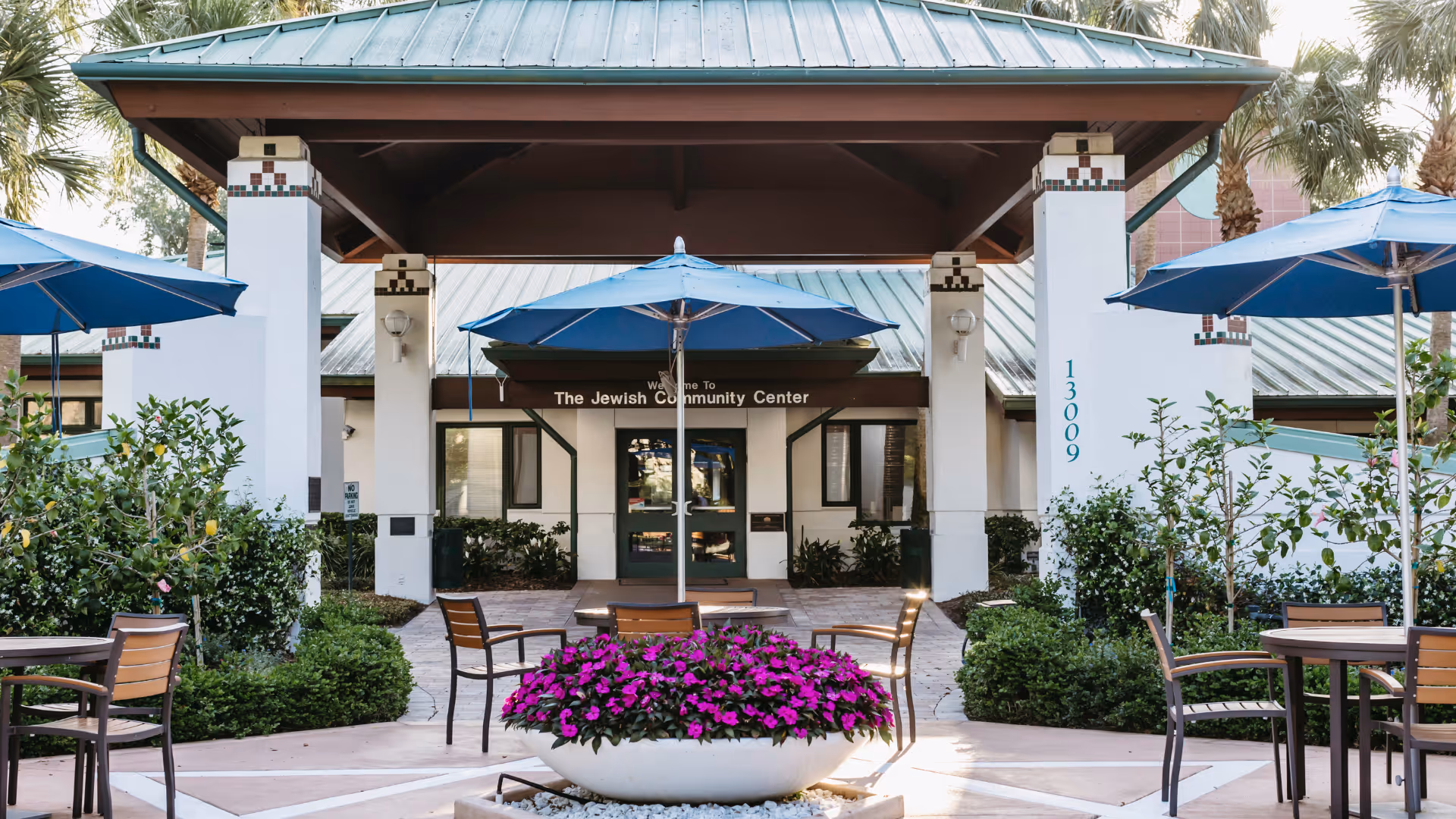 Outdoor seating area with tables, chairs, and blue umbrellas in front of a building entrance labeled 'The Jewish Community Center'. The area is surrounded by greenery and flowers, including a large planter with vibrant purple flowers in the center.