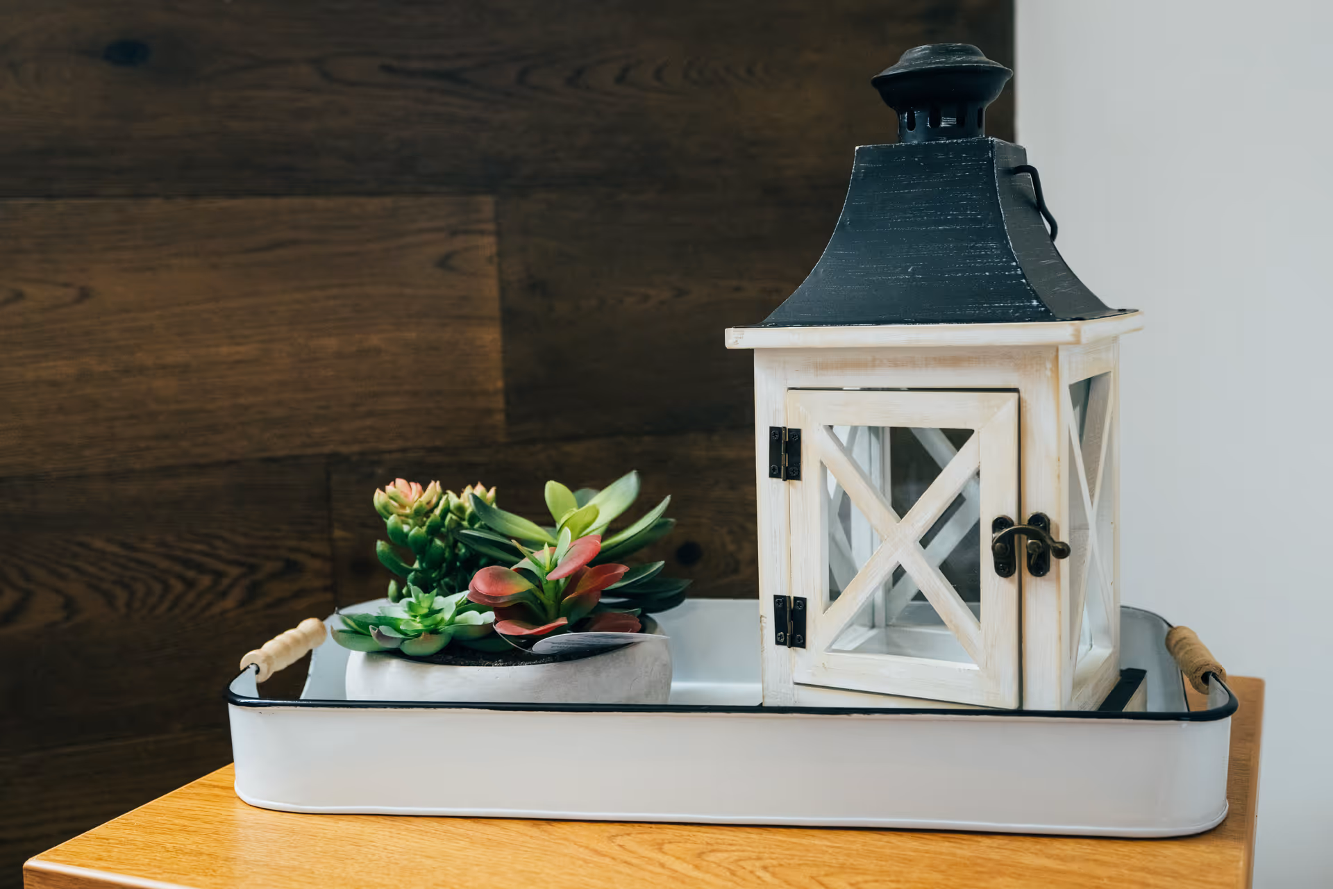 A white tray with wooden handles on a wooden table, holding a small white decorative lantern with a black top and a small pot of green and red succulent plants, set against a wooden paneled wall and a white wall.