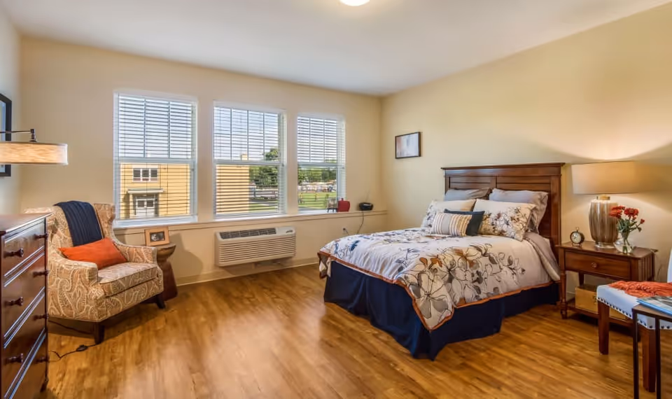 Sunlit furnished bedroom with a bed, armchair, dresser, and large windows.