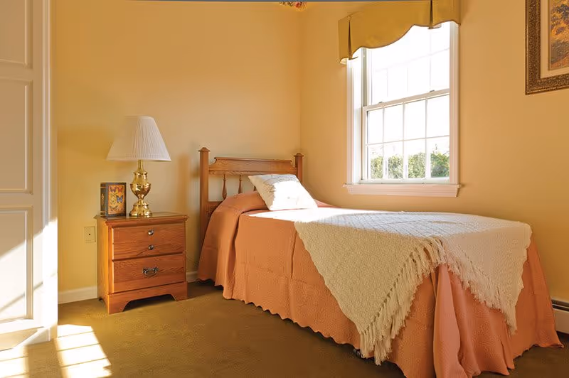A small bedroom with a single wooden bed covered with a peach bedspread and a white knitted throw. There is a wooden nightstand with a brass lamp and a framed picture next to the bed. A window with a yellow valance lets in natural light, illuminating the room with soft yellow walls and a carpeted floor.