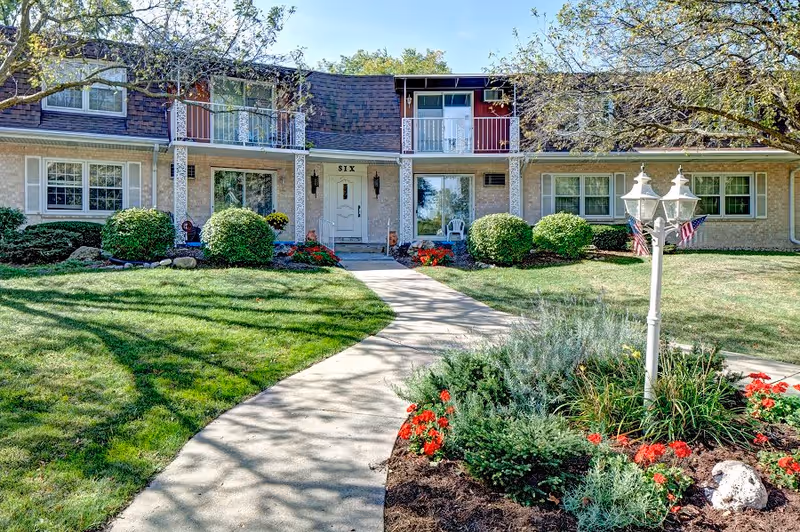 Front exterior view of Whitcomb Square Apartments showing a two-story building with beige brick walls, white-framed windows, and a central white door labeled 'SIX'. A curved concrete pathway leads through a well-maintained lawn and landscaped garden with bushes, flowers, and a white lamp post with American flags. Trees provide partial shade over the scene.