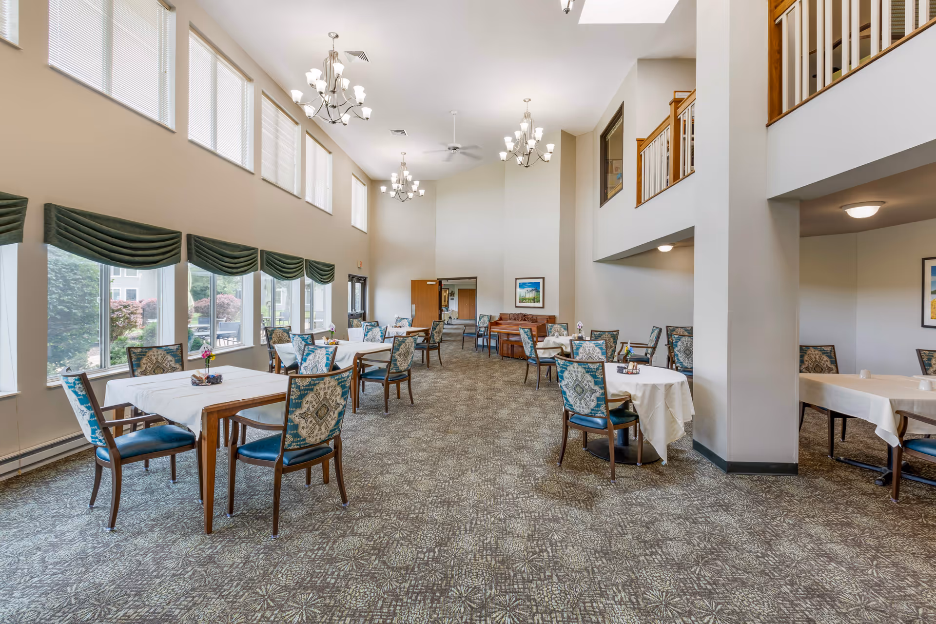 A spacious senior living dining room with multiple tables covered in white tablecloths and surrounded by patterned chairs. Large windows on the left side let in natural light, and green valances hang above them. The room features high ceilings with chandeliers and a carpeted floor with a geometric pattern. There is a piano against the far wall and a second-floor balcony overlooking the area.