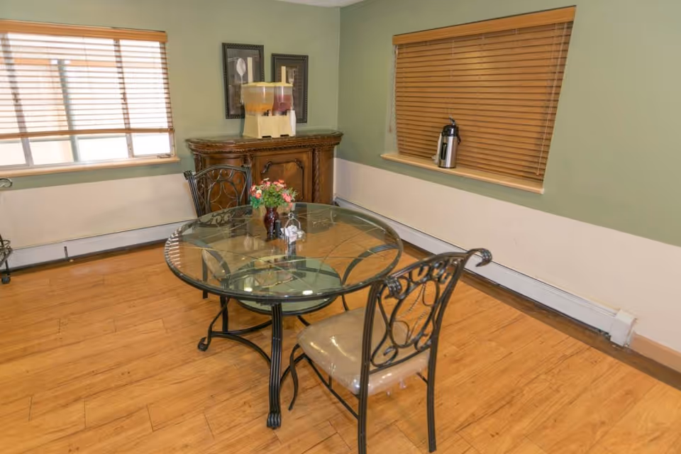 A small dining area with a round glass table and three metal chairs with cushioned seats. The room has light green walls with a white lower section, wooden blinds on two windows, and a wooden floor. A wooden cabinet in the corner holds a beverage dispenser and two framed pictures hang above it. A coffee thermos is placed on the windowsill.
