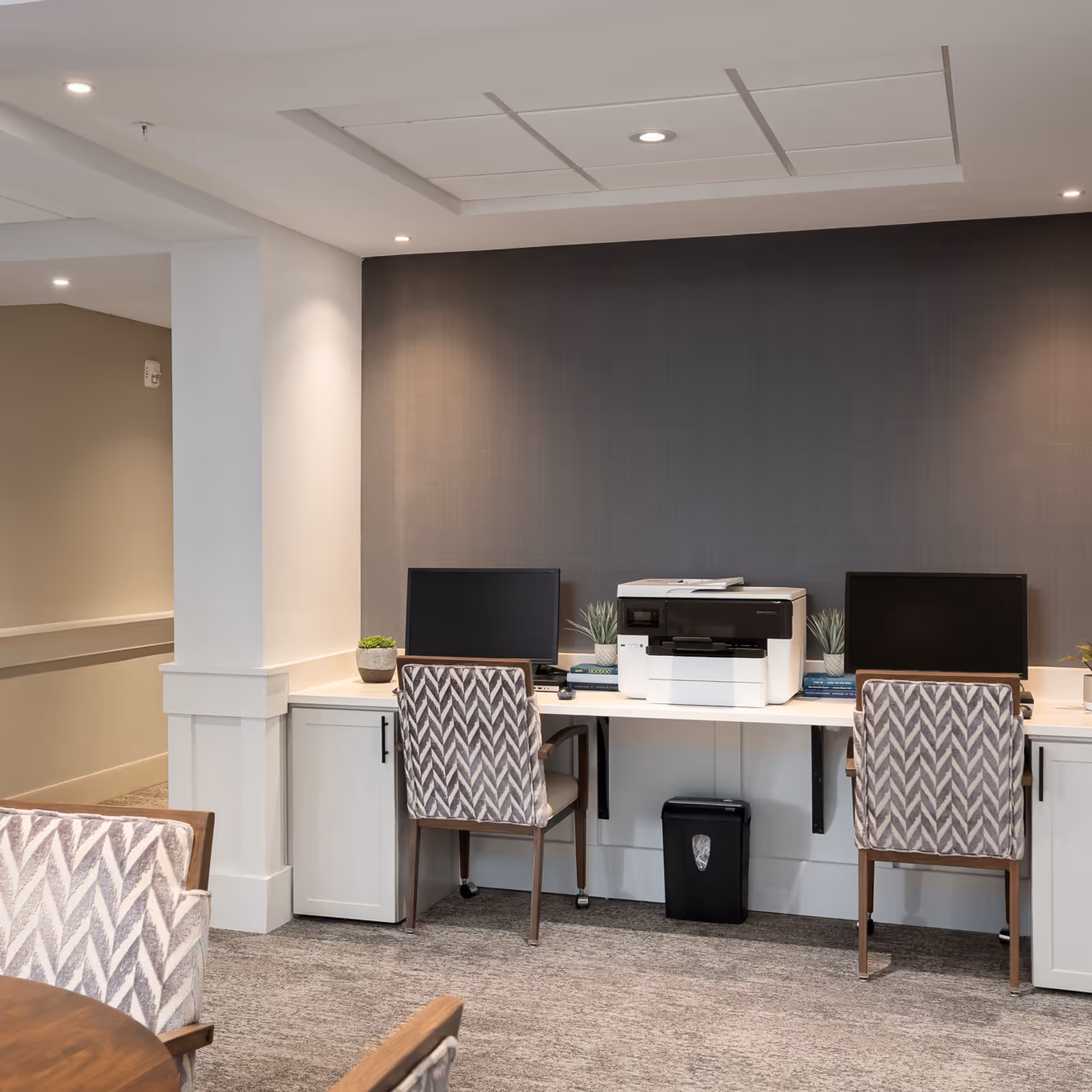 A modern office or computer workstation area with two computer monitors, a printer in the center, two patterned chairs, small potted plants, and a trash bin underneath the desk. The room has a gray accent wall and recessed ceiling lights.