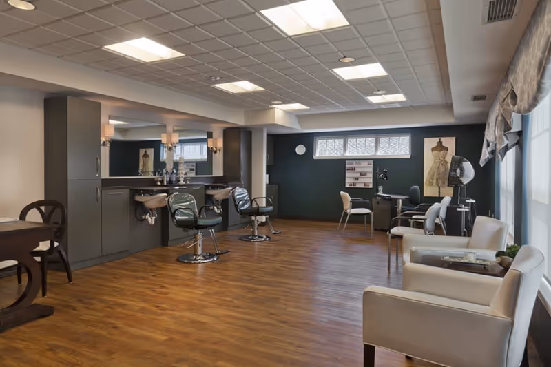 A spacious and well-lit salon area in an assisted living facility with wooden flooring, two salon chairs in front of mirrors and sinks, a manicure station with chairs, and comfortable armchairs near large windows with curtains.
