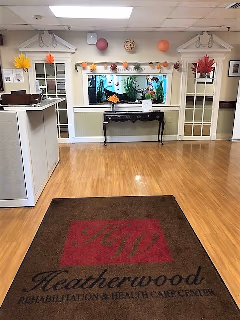 Interior view of a reception or lobby area at Heatherwood Rehabilitation & Health Care Center featuring a wooden floor, a large brown and red mat with the facility's name and logo, a decorative black table with a flower vase, a large fish tank embedded in the wall, and two glass-paneled doors on either side. The walls are decorated with autumn-themed paper leaves and hanging paper lanterns.