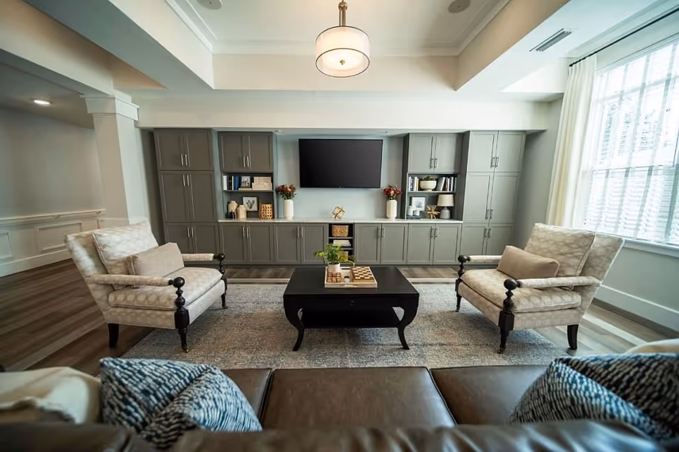Sunlit residential living room with built-in cabinetry and a wall-mounted TV, two upholstered armchairs and a central coffee table.