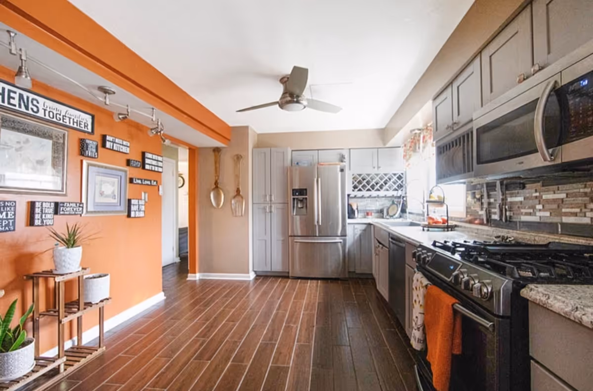 Bright modern kitchen with stainless steel appliances, gray cabinets, wood-look tile floor and an orange accent wall decorated with framed signs.