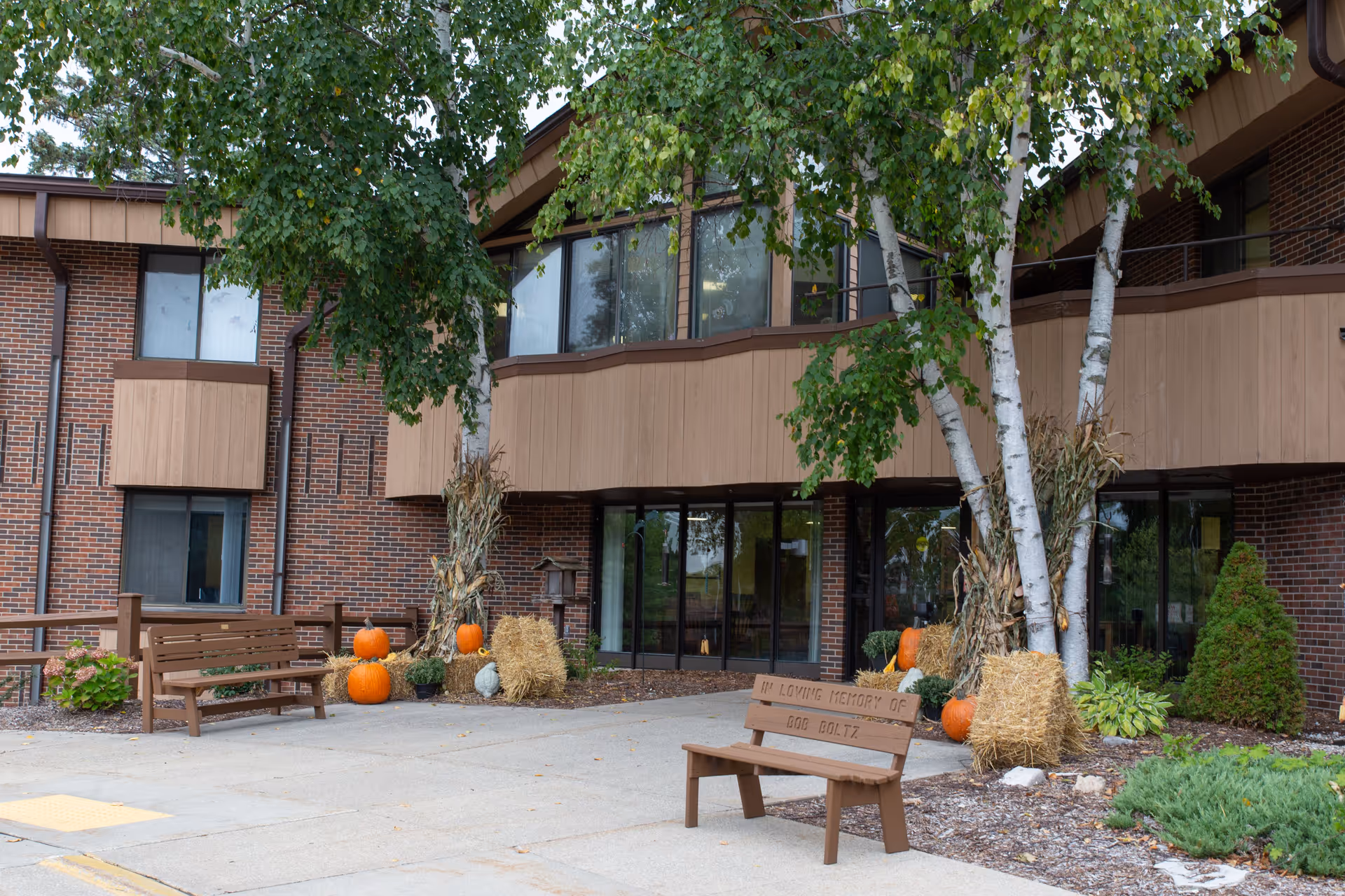 Exterior view of a brick building with large windows and wooden accents, featuring two wooden benches on a concrete walkway. One bench has a memorial plaque. The area is decorated with pumpkins, hay bales, and cornstalks, with trees and shrubs nearby.