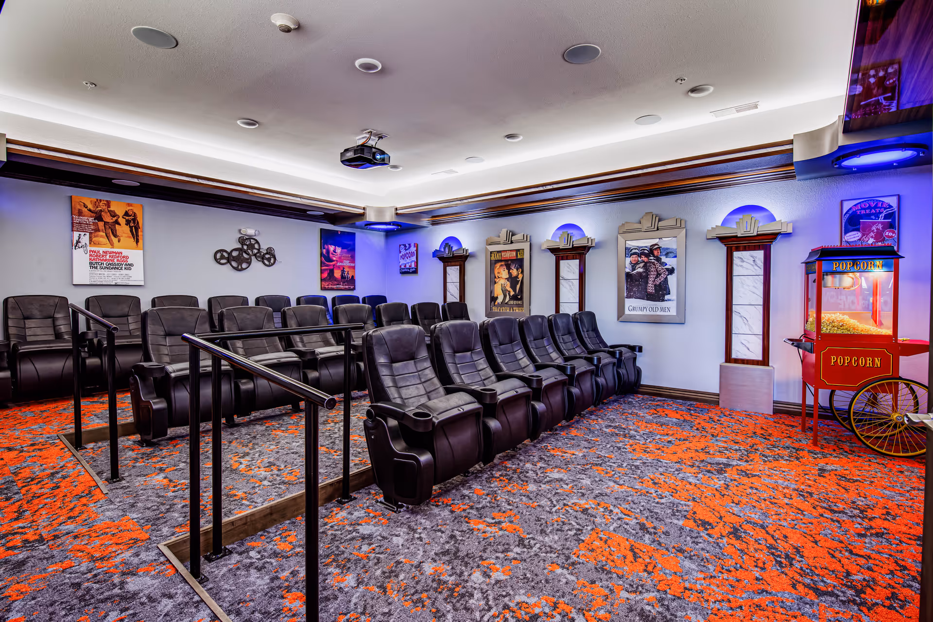 A small movie theater room with two rows of black leather reclining chairs arranged on a carpeted floor with a gray and orange pattern. The walls are decorated with vintage movie posters and decorative columns with blue accent lighting. A red popcorn machine is positioned in the corner of the room. A ceiling-mounted projector is visible above the seating area.