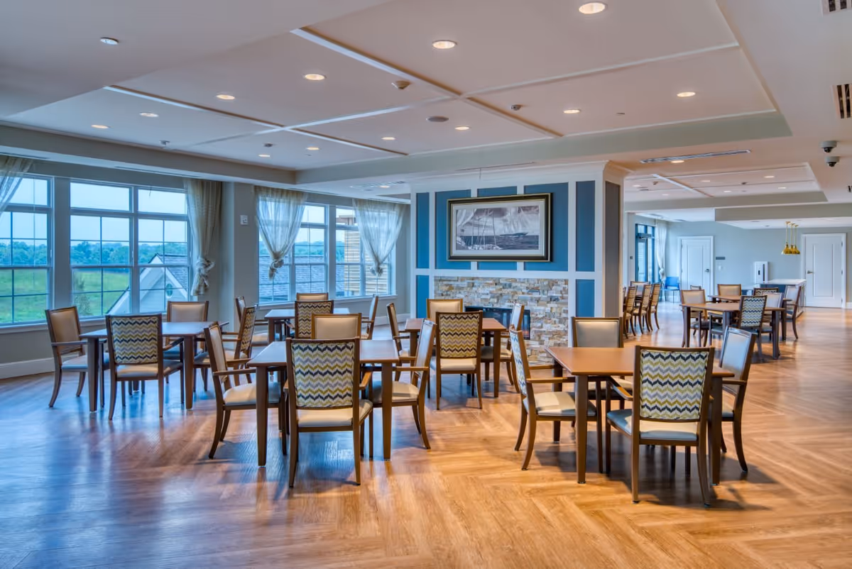 A spacious dining room with multiple wooden tables and chairs arranged neatly. Large windows with sheer curtains allow natural light to fill the room. The walls feature a decorative blue panel with a framed picture above a stone fireplace. The floor is wooden with a herringbone pattern, and the ceiling has recessed lighting.