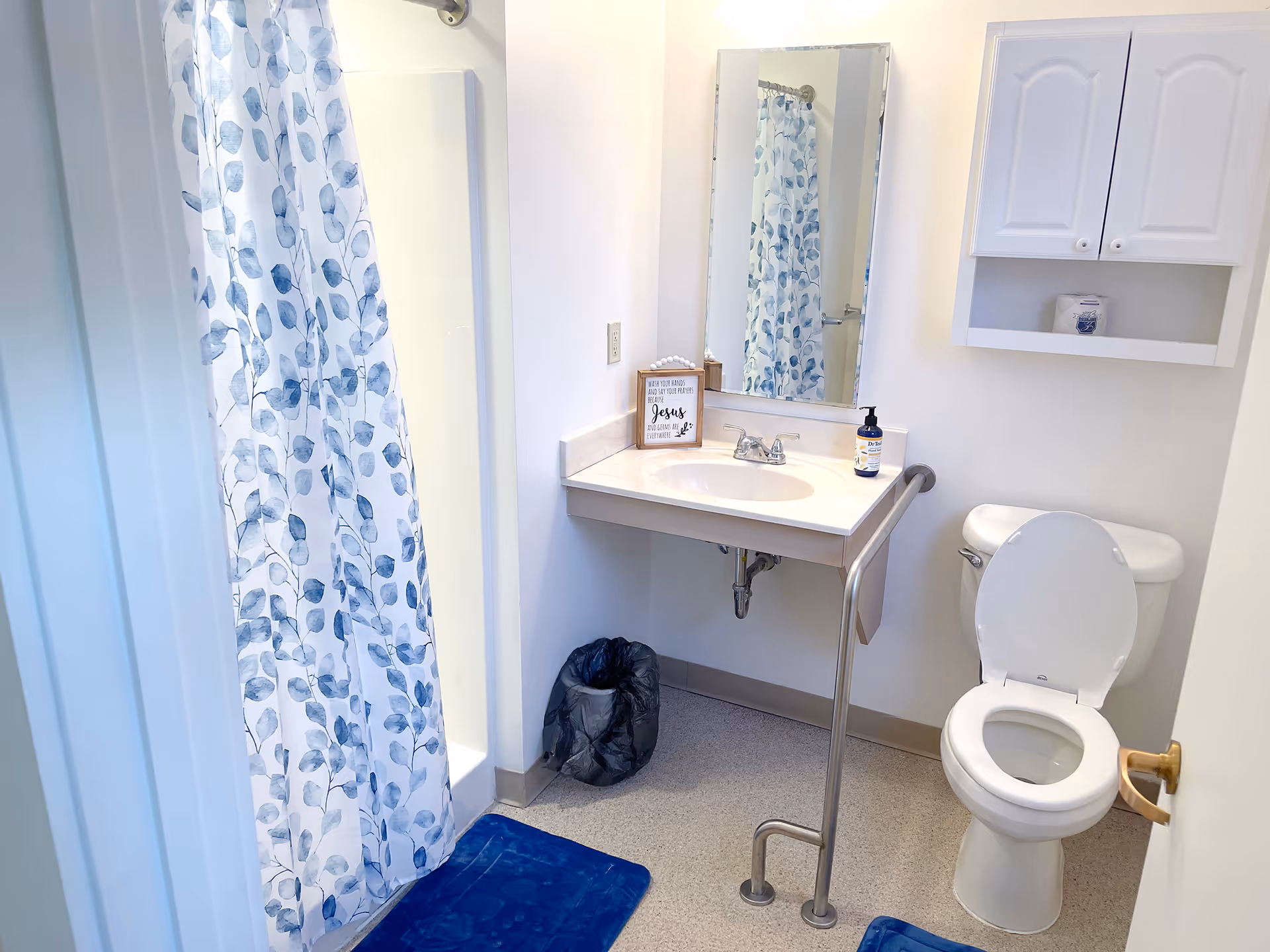A clean bathroom with a white toilet, a sink with a mirror above it, a shower with a blue and white leaf-patterned curtain, a blue bath mat on the floor, a small trash can, and a white wall cabinet. There is also a metal grab bar next to the toilet and sink for support.