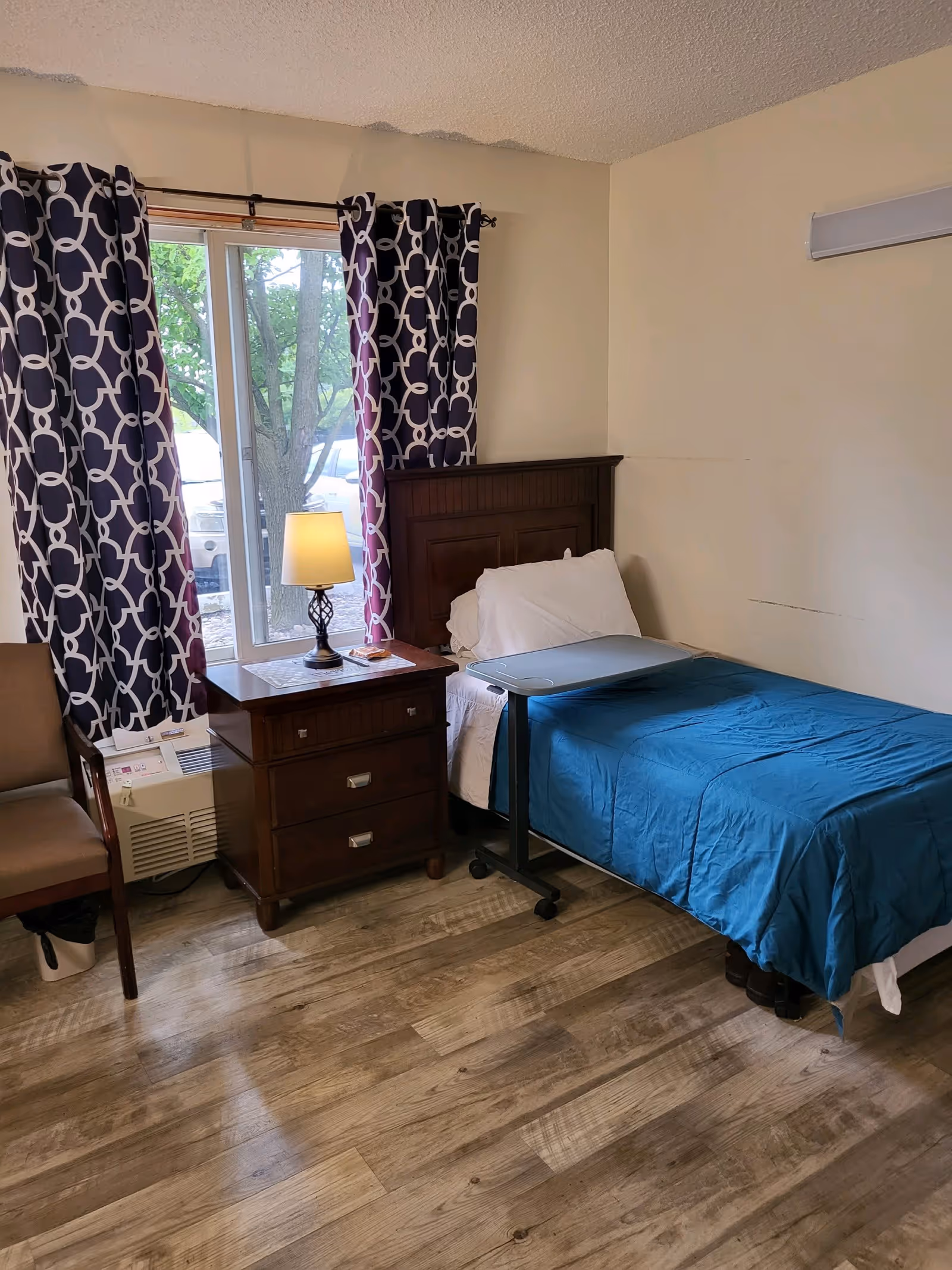 A small bedroom with a single bed covered with a blue blanket and white pillows. Next to the bed is a dark wooden nightstand with three drawers, a lamp, and some items on top. A window with patterned curtains is behind the nightstand. There is a chair and an air conditioning unit near the window. The floor is wooden, and the walls are plain light-colored.
