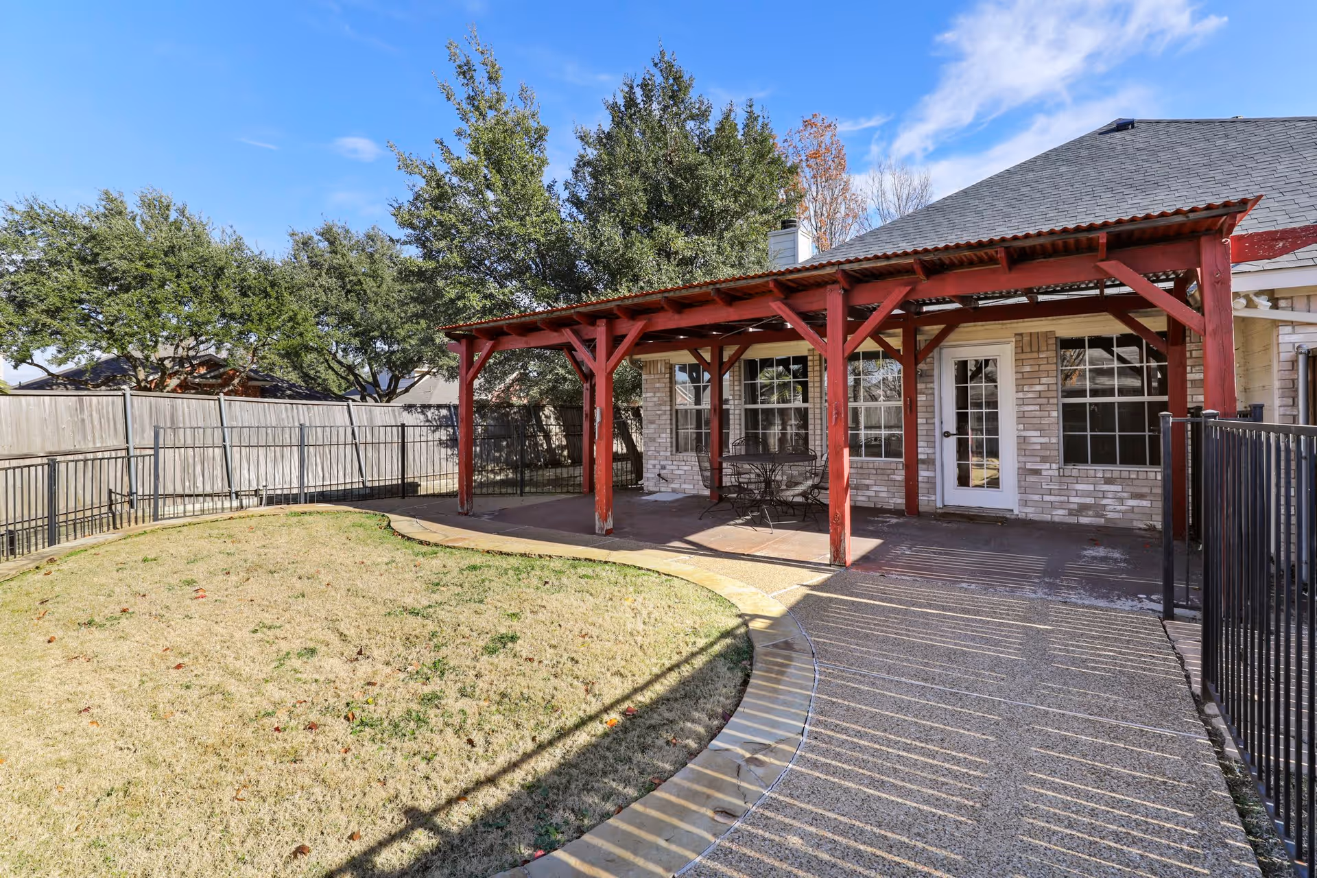 Outdoor patio area with a red wooden pergola attached to a brick building. The patio has a small round table with chairs underneath the pergola. There is a curved concrete pathway leading to the patio, surrounded by a grassy lawn and fenced yard with trees in the background under a blue sky.