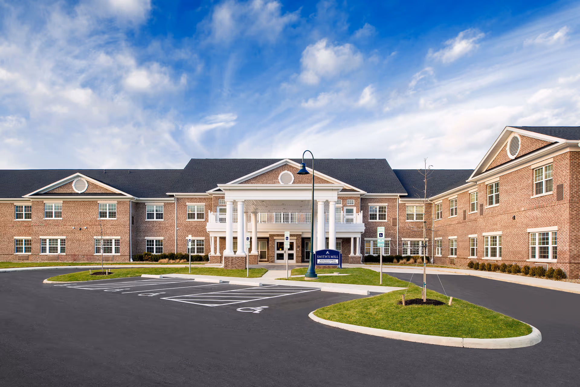 Front exterior view of Smith's Mill Health Campus, a large brick building with white columns at the entrance, a blue sky with some clouds, and a parking area with handicap parking spaces in front.