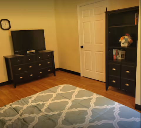 A bedroom with a bed covered in a blue and white patterned comforter in the foreground. Against the walls are a dark wooden dresser with a flat-screen TV on top and a matching dark wooden cabinet with shelves containing books and a flower arrangement. A white door is visible in the corner of the room.