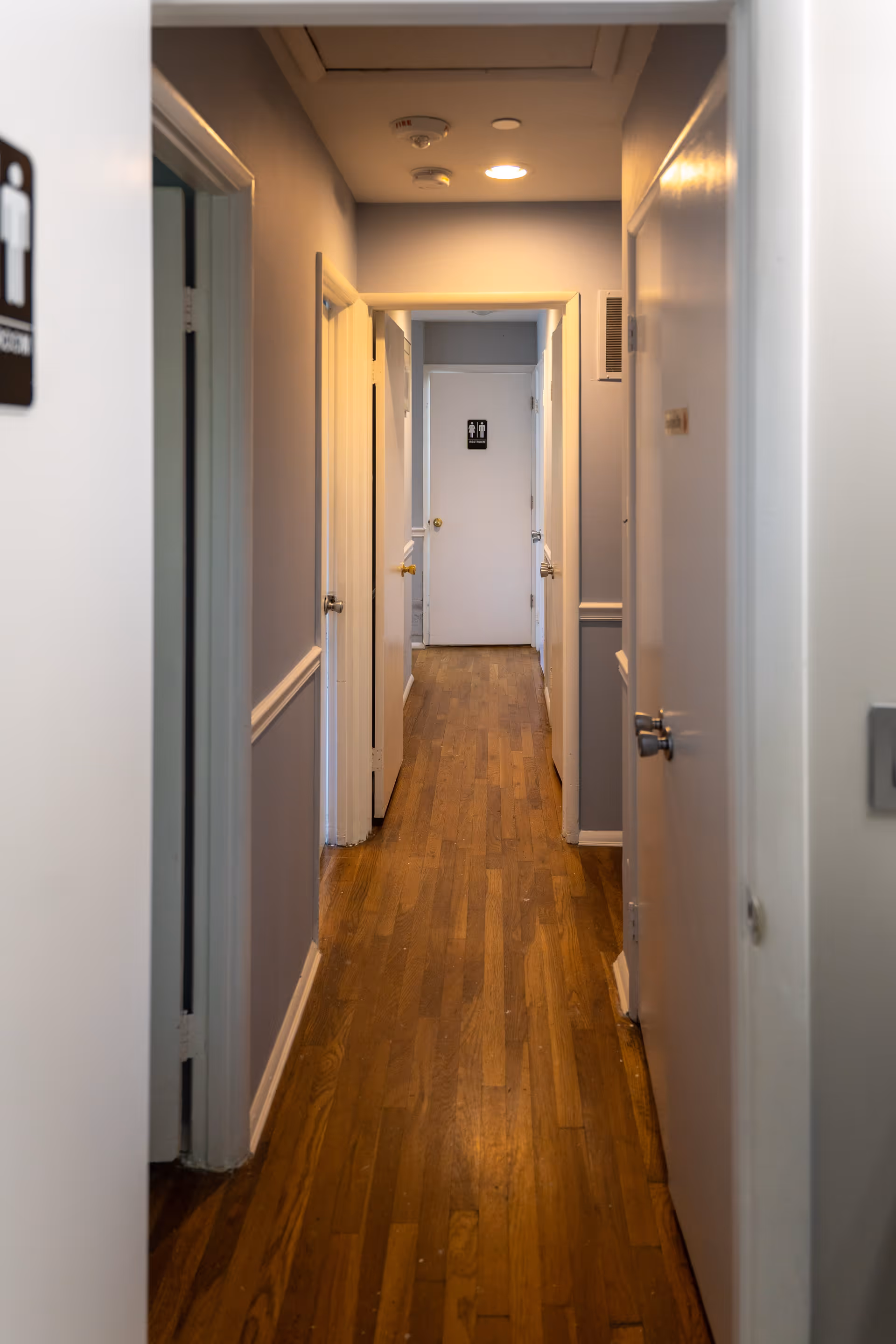 Narrow interior hallway with wood floors and white doors leading to a restroom door at the far end.