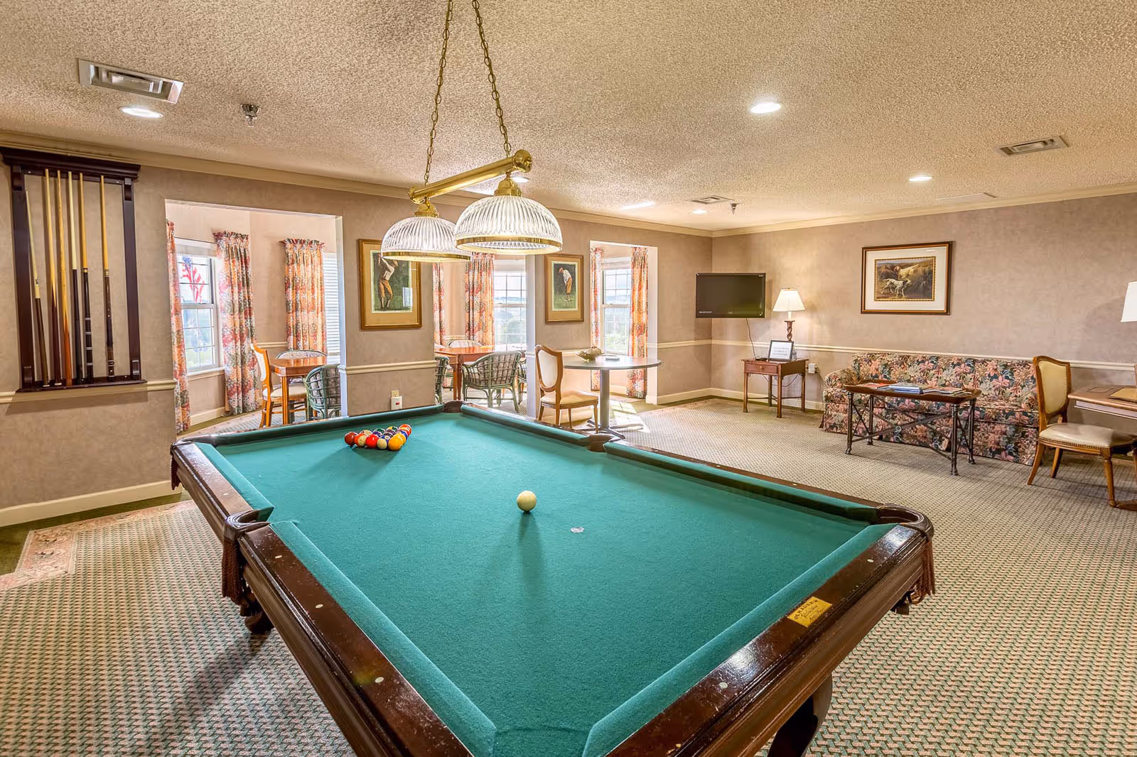 A recreational room with a green felt pool table in the center, a rack of pool cues mounted on the wall, and a set of billiard balls arranged on the table. The room has beige walls, patterned carpet, and ceiling lights. In the background, there is a seating area with a floral couch, a coffee table, a wall-mounted TV, and framed artwork. There are also two small tables with chairs near windows with floral curtains.