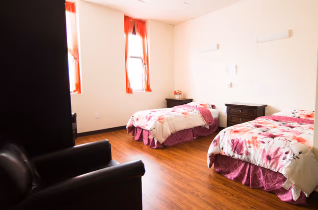 A bright bedroom with two twin beds covered in floral bedspreads with pink and purple accents. There are two windows with orange curtains letting in natural light. The room has wooden flooring, a dark armchair in the foreground, and two dark wooden nightstands between and beside the beds.