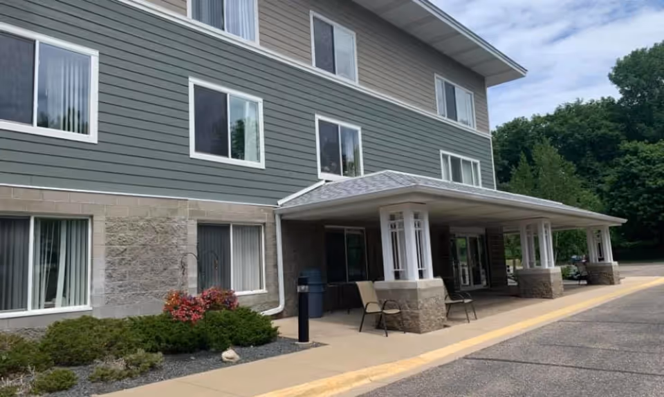 Exterior view of a multi-story senior living facility building with gray siding and stone accents. The entrance has a covered porch supported by white columns, with chairs placed outside. There are windows on all visible floors and some greenery and flowers near the building.