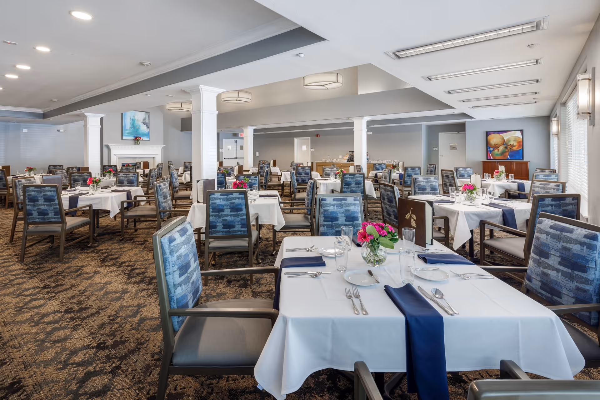 A spacious dining room with multiple tables covered in white tablecloths and navy blue napkins. Each table is set with plates, glasses, silverware, and a small vase of pink flowers. The room features blue patterned chairs, carpeted flooring, white walls, and ceiling lights. There is a fireplace with a painting above it on one side and another colorful painting on the far wall.