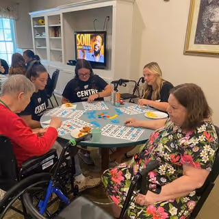 Five people sitting around a round table playing bingo in a well-lit room. Two women in wheelchairs are visible, and three other individuals are seated. A television mounted on the wall shows a program titled 'SOUL'. Shelves with books and decorative items are in the background.